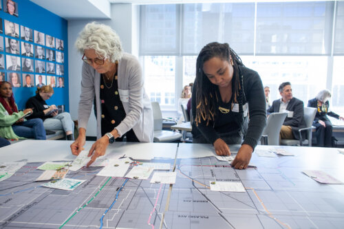 two people reach over a table. on the left is a light skinned person with white hair. on the right is a Black woman with black hair. They are arranging papers on a diagram that is placed on a table. Behind them, people are reading and discussing.