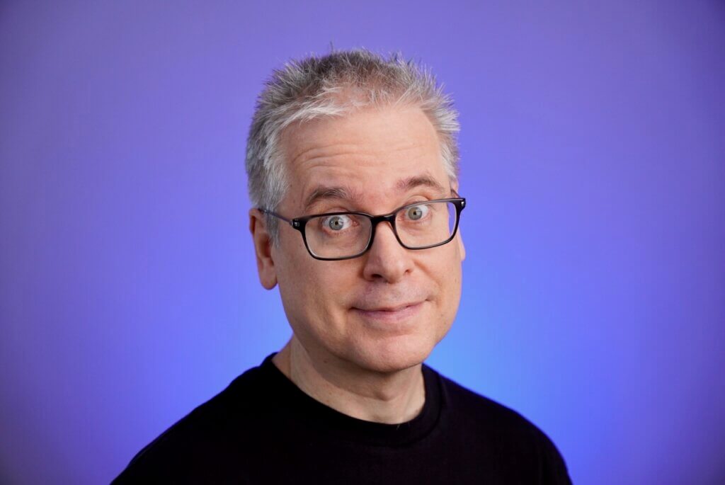 a headshot of a white man with short white and gray hair and black glasses. He is wearing a plain black shirt and there is an indigo background.