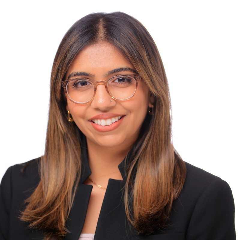 Person with light brown skin and brown hair smiles at the camera, wearing glasses and a black shirt over a white shirt.